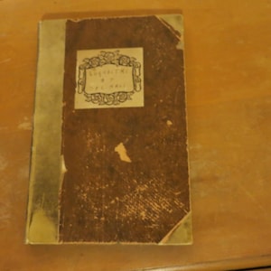 May include: An antique book with a worn, brown cover and a gold spine. The cover features a decorative label with Japanese text. The book is placed on a wooden surface.