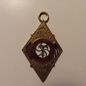 May include: A vintage gold-toned diamond-shaped pendant with the text "Toronto Third Triennial Brotherhood Convention 1922." The pendant features a central emblem and flags of the United Kingdom and the United States.