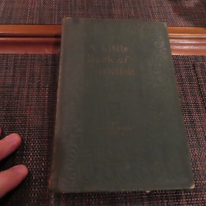 May include: A Little Book of Thoughts, a vintage book with a dark green cover and gold lettering. The book's title is prominently displayed, with decorative floral patterns. The book is slightly worn, suggesting age and use.
