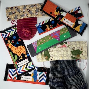 Puede incluir: Una colección de artículos de tela coloridos, que incluyen muñequeras estampadas, una pequeña bolsa de hilo con lana roja y calentadores de piernas a rayas. Las muñequeras presentan varios diseños, como llamas, patrones geométricos y monos calcetín.
