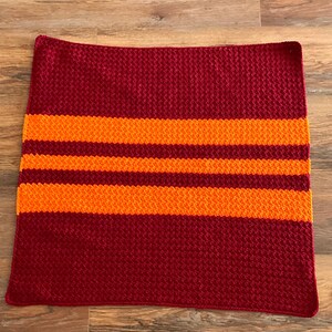 May include: A crocheted blanket with three orange stripes on a maroon background.