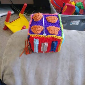 May include: A vibrant, handmade crochet cube toy with diverse textures and elements. The cube features a red base with three zip closures, a purple top with four orange textured shapes, and yellow, pink, and blue edging. Designed for sensory play.