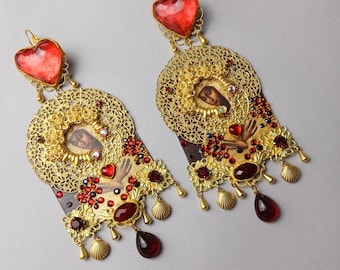 May include: Ornate gold-tone earrings with a heart-shaped red stone at the top. The earrings feature a filigree design, a small image, and red and gold embellishments, including teardrop-shaped dangles and shell charms. The earrings are on a light gray background.