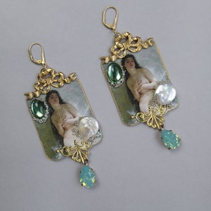 May include: A pair of gold tone earrings with a vintage aesthetic. Each earring features a rectangular pendant with a painted image of a woman, a green gemstone, and a white pearl. The pendants are attached to a gold tone chain with a teardrop-shaped green gemstone.