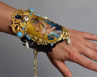 May include: A gold cuff bracelet with a black lace base and a gold-framed image of a mother and child. The bracelet is adorned with turquoise and white pearl accents.