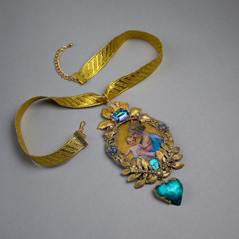 May include: Gold necklace with a large pendant featuring a framed image of the Virgin Mary and baby Jesus. The pendant is decorated with gold leaves, blue and green gemstones, and a heart-shaped charm.
