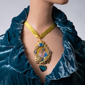 May include: A gold necklace with a pendant featuring a depiction of the Virgin Mary holding a baby Jesus. The pendant is surrounded by gold leaves and blue gemstones. The necklace is worn by a person wearing a teal velvet robe.