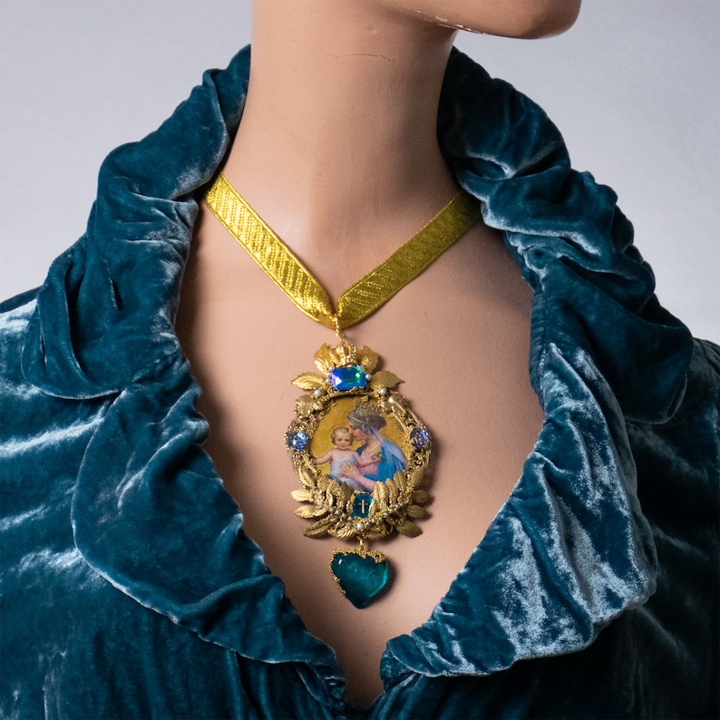 May include: A gold necklace with a pendant featuring a depiction of the Virgin Mary holding a baby Jesus. The pendant is surrounded by gold leaves and blue gemstones. The necklace is worn by a person wearing a teal velvet robe.