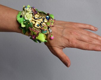 May include: A gold-toned bracelet with a floral design. The bracelet is adorned with green leaves, pink flowers, and colorful gems.