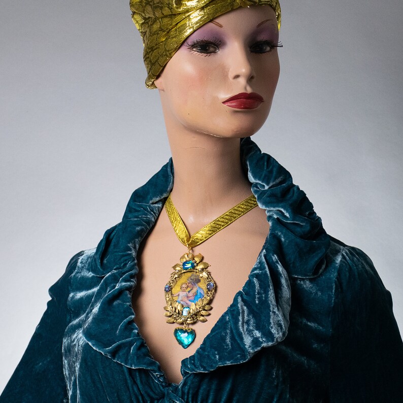 May include: A woman wearing a gold turban and a teal velvet robe with a gold chain necklace featuring a blue jeweled pendant and a gold locket with a painted image of two people.