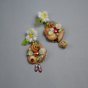 Puede incluir: Un par de pendientes hechos a mano con un grupo de flores blancas, formas marrones parecidas a pasteles y detalles rojos y dorados. Los pendientes están unidos a una cadena dorada con un pequeño dije.