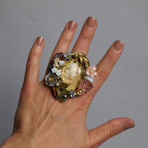 Peut inclure: Une grande bague statement avec un camée représentant le visage d'une femme entouré d'un groupe de fausses fleurs, de perles et de strass. La bague est en métal doré et a un style vintage.