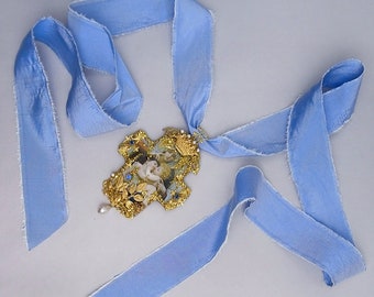 May include: A blue satin ribbon tied around a gold cross pendant with a pearl drop and blue embellishments. The cross features a painted image of angels.
