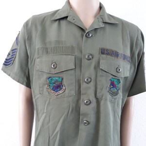 May include: Olive green short-sleeve shirt with two chest pockets and button closures. Features a "U.S. AIR FORCE" patch and two embroidered patches with colorful designs. Includes blue stripes on the left sleeve.