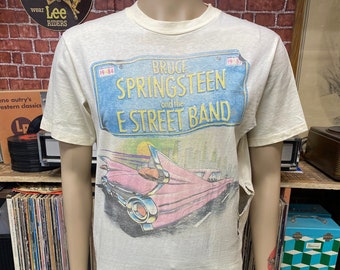 May include: A vintage white t-shirt with a faded image of a pink car and the text "Bruce Springsteen and the E Street Band" on a blue license plate. The license plate has the numbers "19-84" on it.