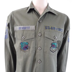 May include: Olive green U.S. Air Force shirt with the name "DEHDRTY" above the left pocket. The shirt has two chest pockets, patches, and the words "U.S. AIR FORCE" above the right pocket. The shirt has long sleeves and button closures.