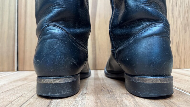 Op de afbeelding: Een paar zwarte leren cowboylaarzen. De laarzen hebben een klassiek ontwerp met een afgeronde hak en een stevige zool. Het leer vertoont wat slijtage, wat bijdraagt aan hun stoere uitstraling.
