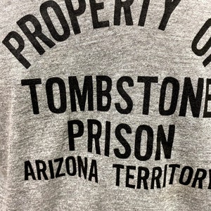 May include: Gray t-shirt with black text that reads "PROPERTY OF TOMBSTONE PRISON ARIZONA TERRITORY".