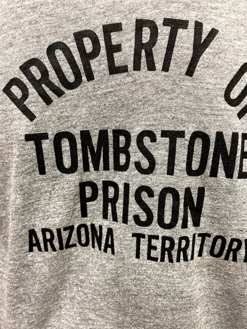 May include: Gray t-shirt with black text that reads "PROPERTY OF TOMBSTONE PRISON ARIZONA TERRITORY".