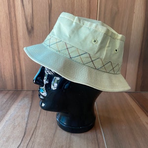 May include: A beige bucket hat with a plaid band around the crown. The hat has two small eyelets on the top.