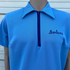 May include: A light blue short-sleeved shirt with a zipper closure and a navy blue collar. The shirt has the name "Darlene" embroidered on the front in navy blue thread.