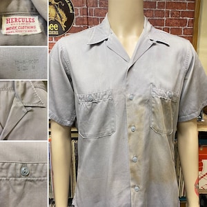 May include: A gray short-sleeved work shirt with a button-down collar and two chest pockets. The shirt has a faded appearance and the label reads "Hercules Work Clothing Sears, Roebuck and Co."