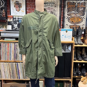 May include: An olive green military-style field parka featuring a high collar, a full-length front zipper concealed by a snap-button placket, long sleeves with adjustable cuffs, and two large flap pockets positioned at the waist area.