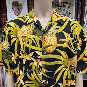 May include: Black button-down shirt with a vibrant pineapple and floral print. The shirt features large yellow pineapples, green leaves, and yellow flowers against a black background. The short-sleeved shirt has a classic collar and wooden buttons.