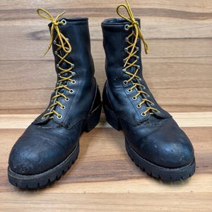 May include: A pair of black leather work boots with yellow laces. The boots have a rugged design with a thick sole and are laced up the front. The boots are set against a wooden background.
