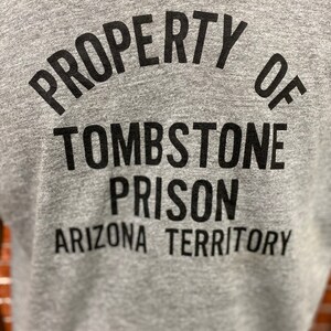 May include: Gray t-shirt with black text that reads "PROPERTY OF TOMBSTONE PRISON ARIZONA TERRITORY".