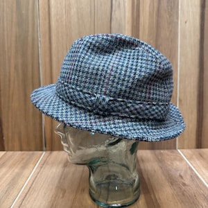 May include: A fedora hat with a houndstooth pattern in shades of blue, green, and red. The hat features a structured brim and a band around the crown. The hat is displayed on a glass head form.