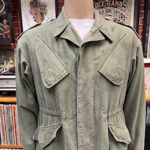 May include: A green military-style jacket with a zipper closure, two large flap pockets on the chest, and two smaller flap pockets on the bottom. The jacket has a drawstring waist and a collar.