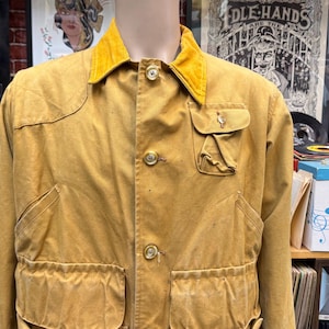 May include: A mustard yellow canvas jacket featuring a contrasting gold corduroy collar, a button-down front with metallic buttons, two large lower patch pockets, and a smaller chest pocket with a flap closure on the left side of the garment.