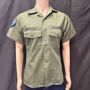 May include: An olive green short-sleeved button-down shirt featuring two chest pockets with flaps. The shirt displays the text MORRISSET above the left pocket, U.S. NAVY above the right pocket, and a circular patch on the right sleeve.