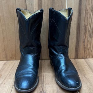 Op de afbeelding: Een paar zwarte leren cowboylaarzen. De laarzen hebben een klassiek ontwerp met een puntige neus en een middelhoge schacht. De laarzen worden gepresenteerd op een houten oppervlak.