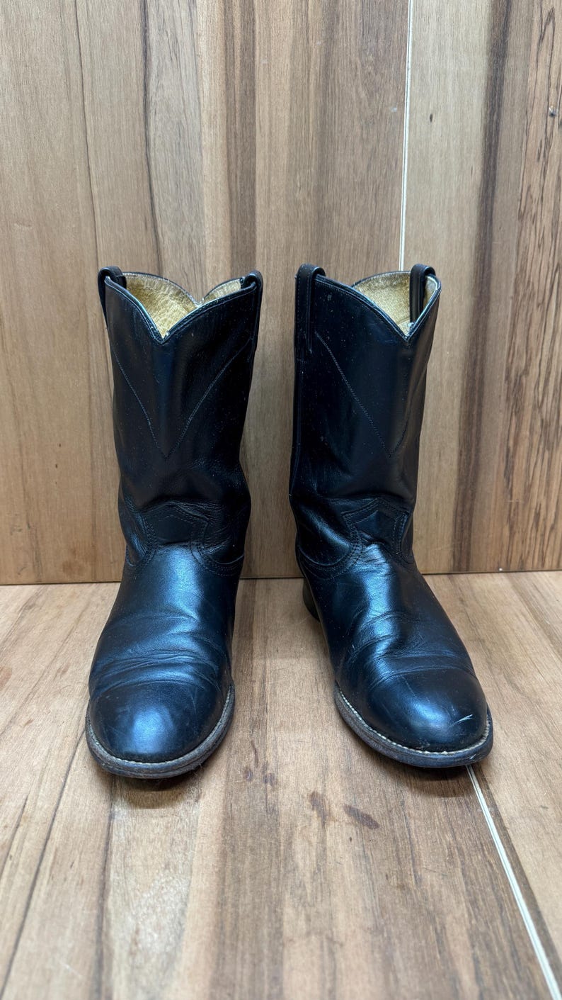 Op de afbeelding: Een paar zwarte leren cowboylaarzen. De laarzen hebben een klassiek ontwerp met een puntige neus en een middelhoge schacht. De laarzen worden gepresenteerd op een houten oppervlak.