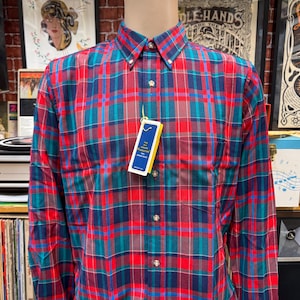 May include: A button-down plaid shirt in red, teal, and navy blue, displayed on a mannequin. The long-sleeved shirt has a classic collar and a chest pocket. A yellow and blue tag hangs from the shirt.