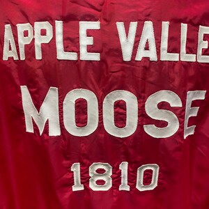 May include: Red jacket with white embroidered text that reads "Apple Valley Moose 1810".