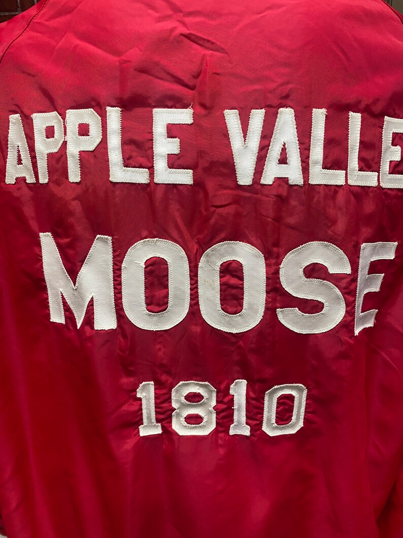 May include: Red jacket with white embroidered text that reads "Apple Valley Moose 1810".