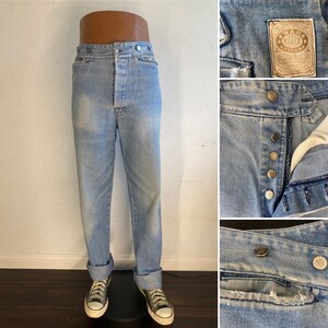 May include: Light blue denim jeans with a button fly and a top stitch detail. The jeans have a faded look and are cuffed at the bottom. The jeans have a label that reads "Wrangler Arizona Top West Size 31 Made in USA".