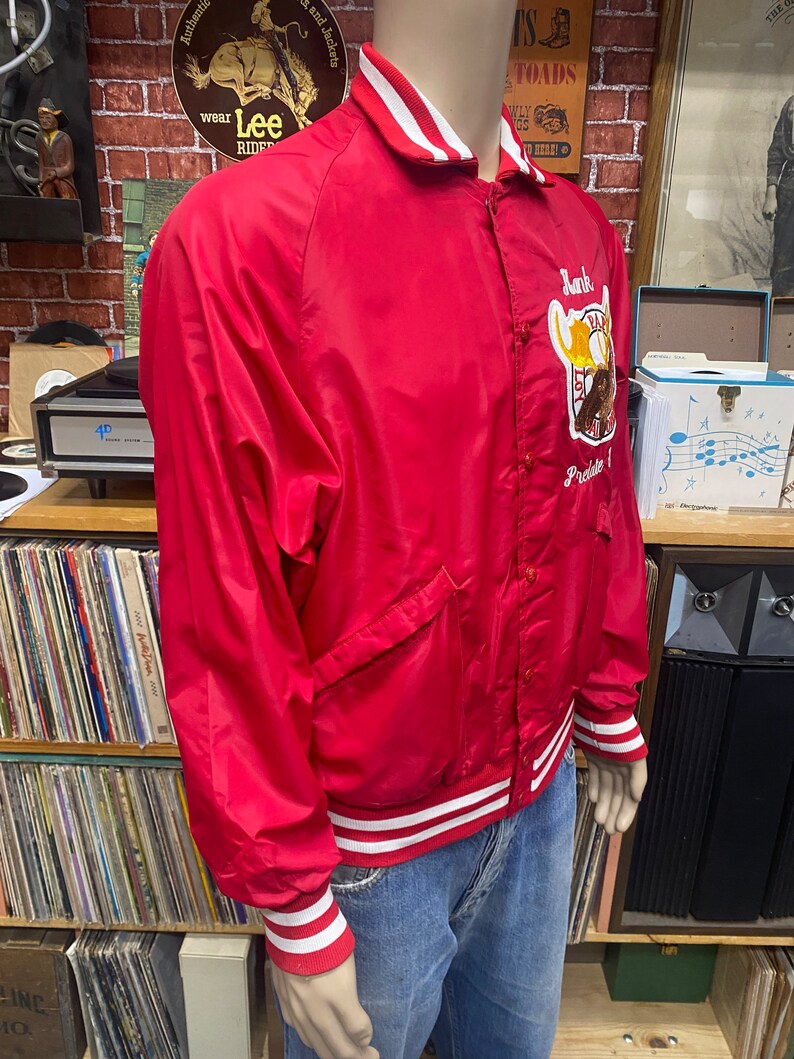 May include: Red satin baseball jacket with white and red striped trim. The jacket has a large embroidered patch on the left side with the text "Lank" and "Prelate".