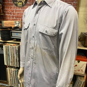 May include: A gray long-sleeved work shirt with two chest pockets and a button-down collar. The shirt has a faded look and is made of a durable fabric.