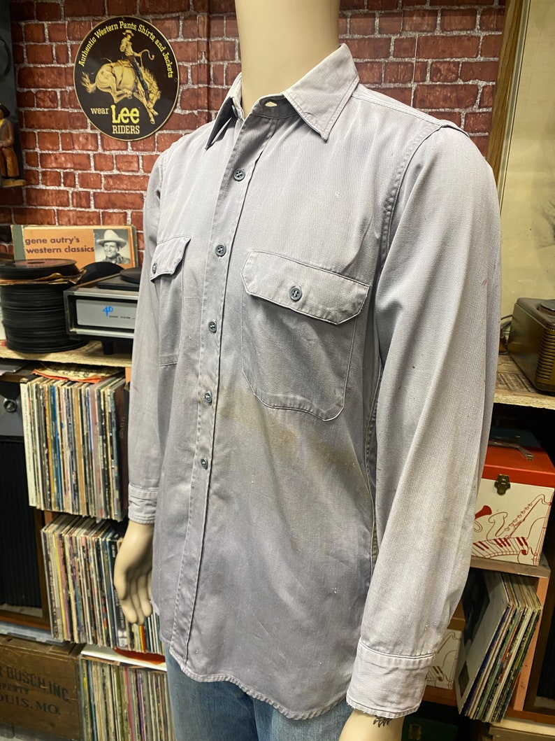 May include: A gray long-sleeved work shirt with two chest pockets and a button-down collar. The shirt has a faded look and is made of a durable fabric.