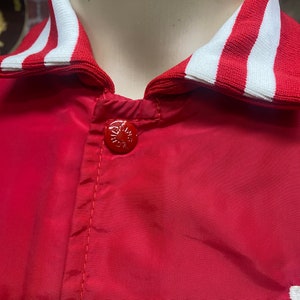 May include: Red satin baseball jacket with a white and red striped collar. The jacket has a single button closure.