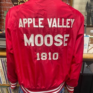 May include: Red satin bomber jacket with white ribbed trim around the collar, cuffs, and waistband. The back of the jacket has white embroidered text that reads "APPLE VALLEY MOOSE 1810".