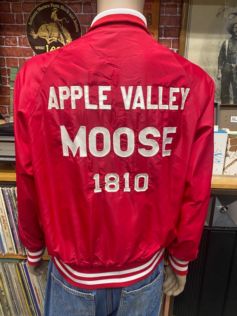 May include: Red satin bomber jacket with white ribbed trim around the collar, cuffs, and waistband. The back of the jacket has white embroidered text that reads "APPLE VALLEY MOOSE 1810".
