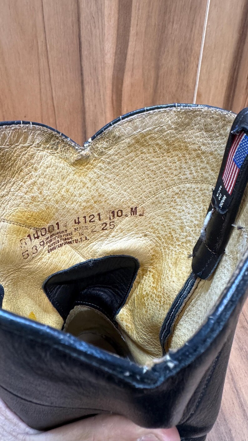 Op de afbeelding: Close-up van de binnenkant van een zwarte leren laars. De binnenkant van de laars is lichtbruin van kleur met zichtbare slijtage. De laars heeft een label met de tekst "MADE IN U.S.A." en andere nummers.