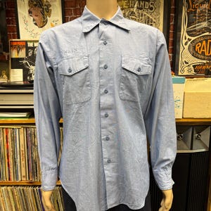 May include: A light blue long-sleeve button-down shirt featuring a pointed collar, two chest pockets with buttoned flaps, and a central button placket. The word SWAIN is embroidered in small, dark blue thread above the right chest pocket.