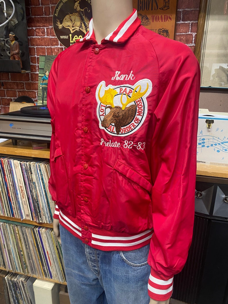 May include: Red satin varsity jacket with white and red striped trim. The jacket has a large embroidered patch with a moose and the text "PAP Loyal Order of Moose Prelate 82-83".
