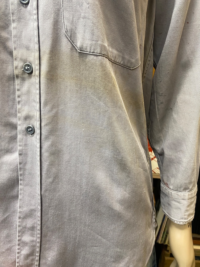 May include: A gray work shirt with a button-up front and a single chest pocket. The shirt has a faded appearance and is made of a durable fabric.